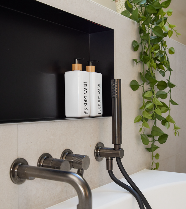 Inbouw nis met flessen douchegel boven bad, voorzien van zwarte achterwand en moderne inbouwkranen voor een strakke look.