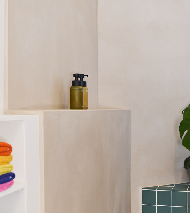 Minimalistische voorzetwand in de badkamer met ingebouwde nis en zeepflessen, naast kleurrijke handdoeken en groene tegels.