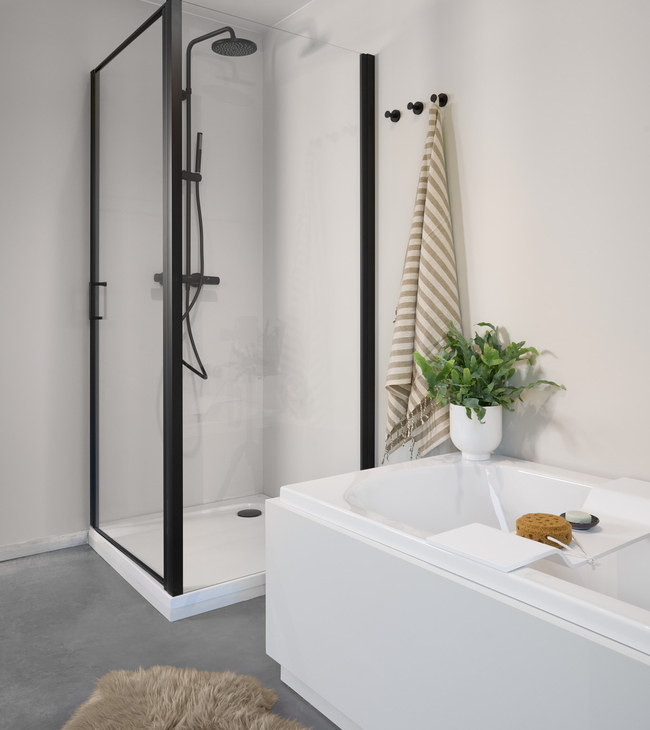 Minimalistische badkamer met zwart doucheprofiel en strak wit bad met omkasting, afgewerkt met groene plant en zachte accessoires.