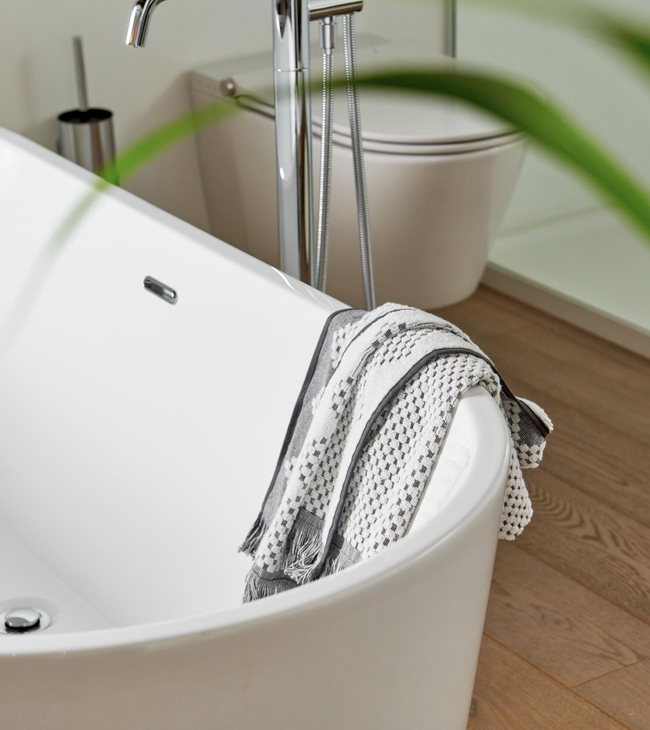 Vrijstaand wit acryl bad met handdoek op houten vloer, gecombineerd met moderne kranen en een minimalistisch interieur.