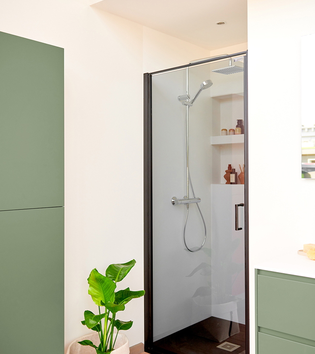 Compacte douche met zwarte deur en witte douchebak in acryl, geplaatst in een frisse badkamer met groene accenten en daglicht.